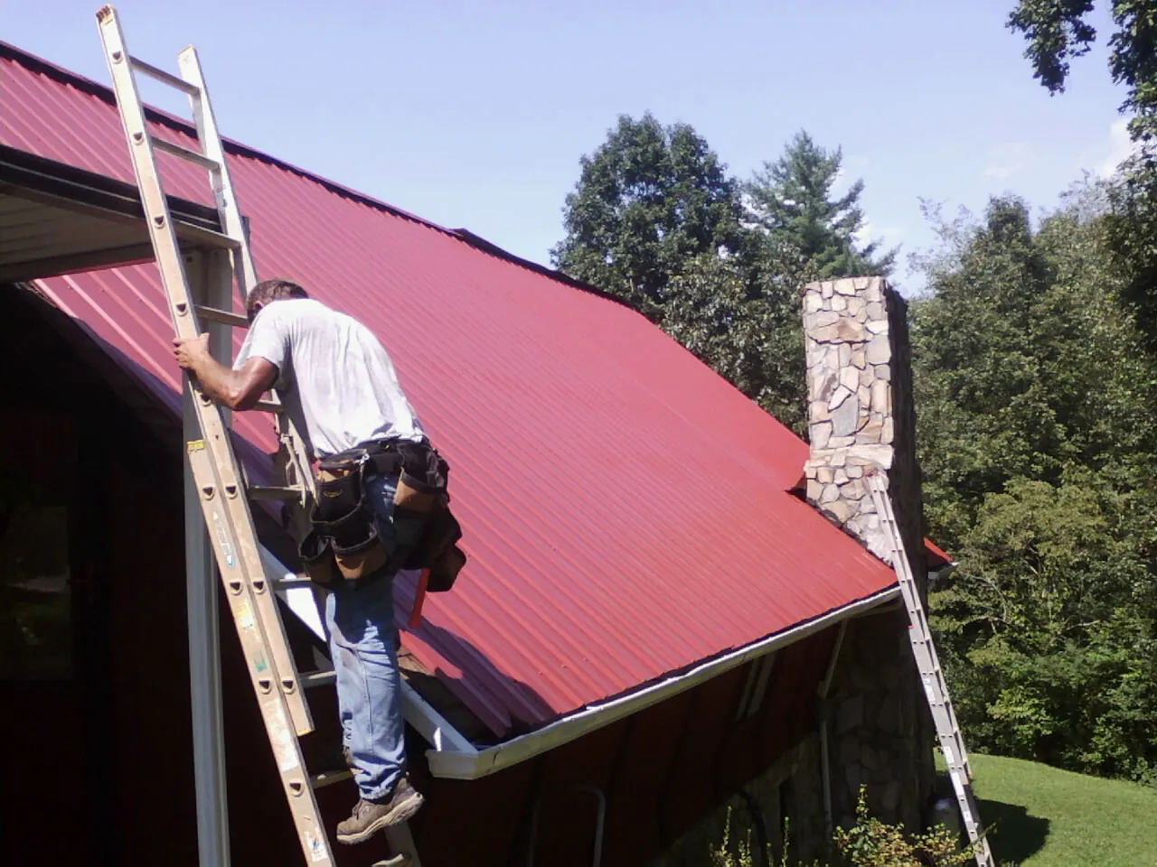Roofer on a ladder beside a red metal roof and stone chimney — Soffit & Fascia Repair by Roofing Brothers in Hinton